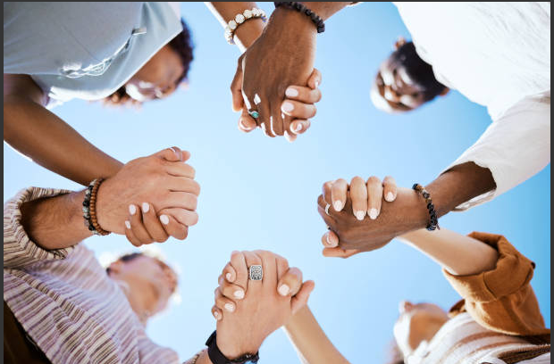 Diverse group of people join hands in a circle against a blue sky, symbolizing teamwork and unity.