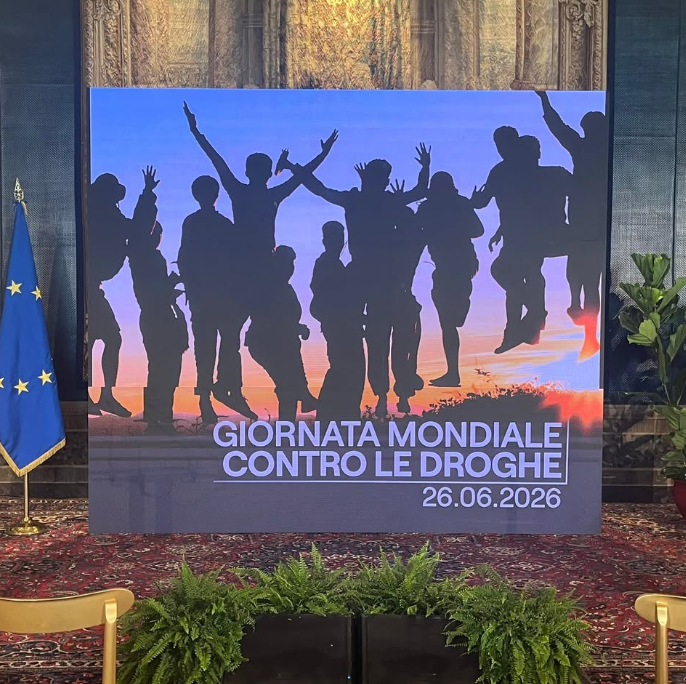 Large display banner with silhouetted group jumping against a sunset, reading 'Giornata Mondiale Contro le Droghe' and the date 26.06.2026; EU flag visible to the left.