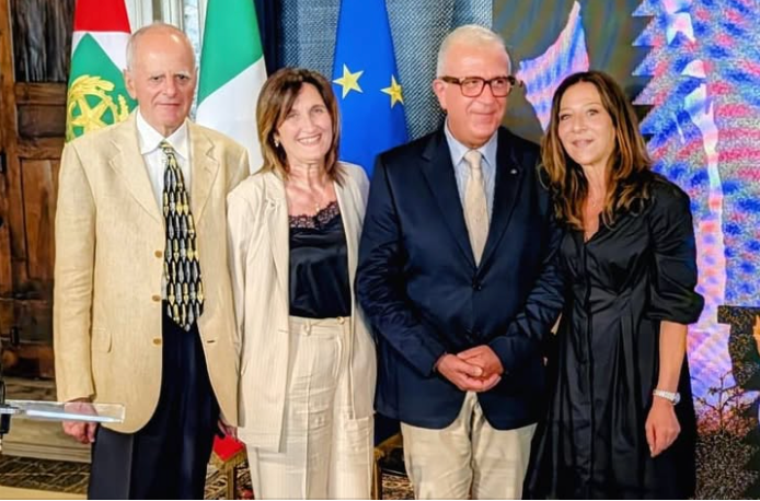 Four professionally dressed individuals posing for a formal group photo, with European Union and national flags in the background.